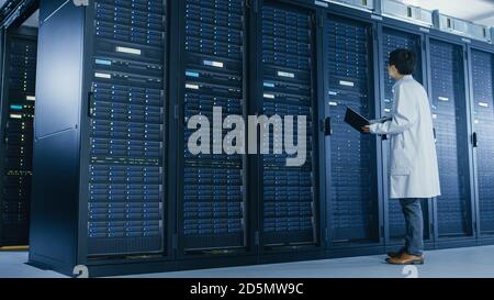 In Data Center: Male IT Specialist Wearing White Coat Stands Beside Server Rack, Uses Laptop Computer to Run Maintenance Diagnostics Tools, Controls Stock Photo