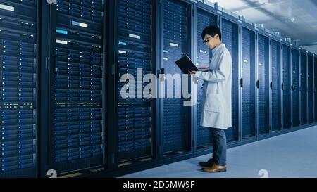 In Data Center: Male IT Specialist Wearing White Coat Stands Beside Server Rack, Uses Laptop Computer to Run Maintenance Diagnostics Tools, Controls Stock Photo