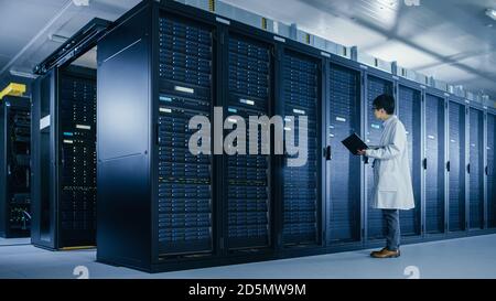 In Data Center: Male IT Specialist Wearing White Coat Stands Beside Server Rack, Uses Laptop Computer to Run Maintenance Diagnostics Tools, Controls Stock Photo