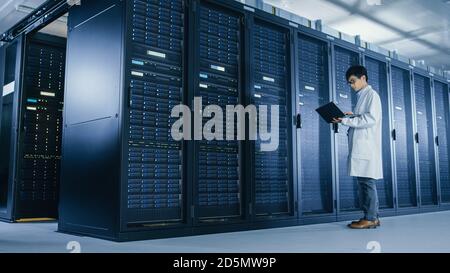 In Data Center: Male IT Specialist Wearing White Coat Stands Beside Server Rack, Uses Laptop Computer to Run Maintenance Diagnostics Tools, Controls Stock Photo