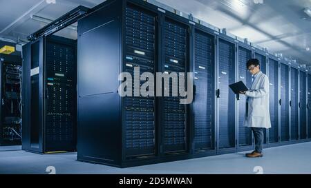 In Data Center: Male IT Specialist Wearing White Coat Stands Beside Server Rack, Uses Laptop Computer to Run Maintenance Diagnostics Tools, Controls Stock Photo
