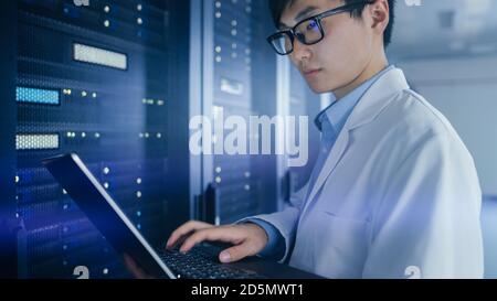 In Data Center: Male IT Specialist Wearing White Coat Stands Beside Server Rack, Uses Laptop Computer to Run Maintenance Diagnostics Tools, Controls Stock Photo