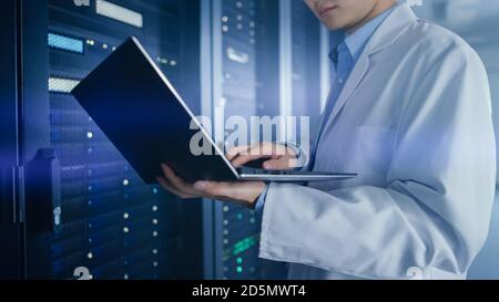 In Data Center: Male IT Specialist Wearing White Coat Stands Beside Server Rack, Uses Laptop Computer to Run Maintenance Diagnostics Tools, Controls Stock Photo