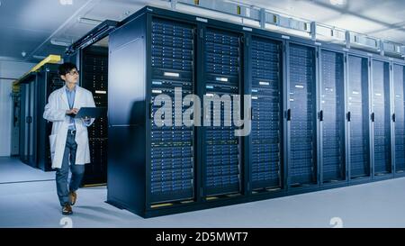 In Data Center: Male IT Specialist Wearing White Coat Walks Alongside Row of Server Racks, Uses Laptop Computer to Run Maintenance Diagnostics Tools Stock Photo
