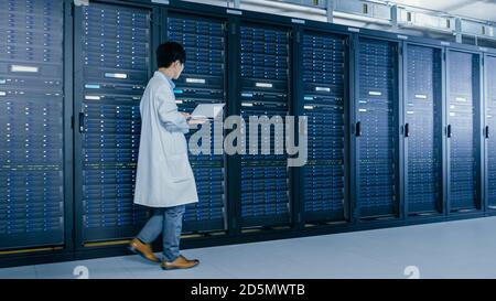 In Data Center: Male IT Specialist Wearing White Coat Walks Alongside Row of Server Racks, Uses Laptop Computer to Run Maintenance Diagnostics Tools Stock Photo