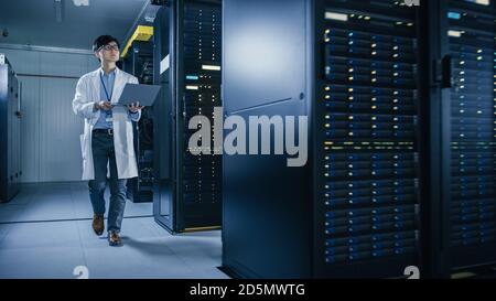 In Data Center: Male IT Technician Wearing White Coat Walking alongside Server Racks, Uses Laptop Computer to Run Maintenance Diagnostics Tools. He is Stock Photo