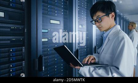 In Data Center: Male and Female IT Specialists Wearing White Coats Work with Server Racks, Use Laptops to Run Maintenance Diagnostics. People wearing Stock Photo