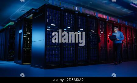 In Data Center: Male IT Specialist Walks along the Row of Operational Server Racks, Uses Laptop for Maintenance. Concept for Telecommunications, Cloud Stock Photo