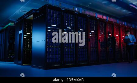 In Data Center: Male IT Specialist Walks along the Row of Operational Server Racks, Uses Laptop for Maintenance. Concept for Telecommunications, Cloud Stock Photo