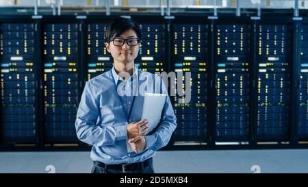 In the Modern Data Center: Portrait of IT Engineer Stands Beside the Row of Server Racks, Finishing Maintenance and Diagnostics Procedure and Holding Stock Photo
