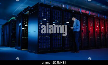 In Data Center: Male IT Specialist Walks along the Row of Operational Server Racks, Uses Laptop for Maintenance. Concept for Telecommunications, Cloud Stock Photo