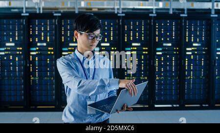 In Dark Data Center: Male IT Specialist Stands Beside Row of Operational Server Racks, Uses Laptop for Maintenance. Concept for Cloud Computing Stock Photo