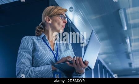 In Data Center: Low-Angle Portrait Shot of a Female IT Technician Running Maintenance Programme on Laptop, Controls Operational Server Rack. Modern Stock Photo