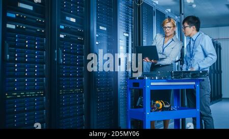 In the Modern Data Center: Engineer and IT Specialist Work with Server Racks, on a Pushcart Equipment for Installing New Hardware. Specialists Doing Stock Photo
