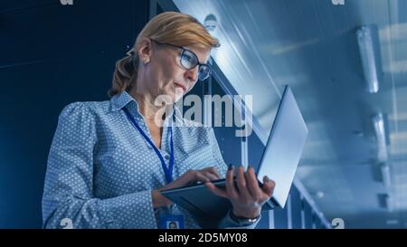 In Data Center: Low-Angle Portrait Shot of a Female IT Technician Running Maintenance Programme on Laptop, Controls Operational Server Rack. Modern Stock Photo
