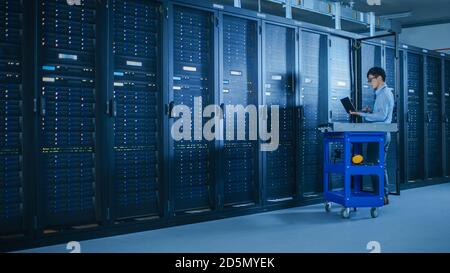 In Data Center: Male IT Technician Running Maintenance Programme on a Laptop, Controls Operational Server Rack Optimal Functioning. Modern High-Tech Stock Photo