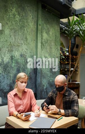 Young people in masks sitting at the table and using their smart phones during meeting in cafe Stock Photo