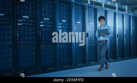 In the Modern Data Center: IT Engineer Working with Server Racks, Uses Laptop for Maintenance and Diagnostics. Checking Stability of the System Stock Photo