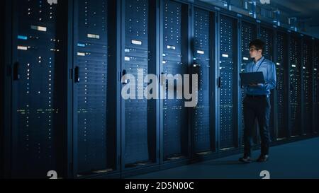 In Dark Data Center: Male IT Specialist Walks along the Row of Operational Server Racks, Uses Laptop for Maintenance. Concept for Cloud Computing Stock Photo