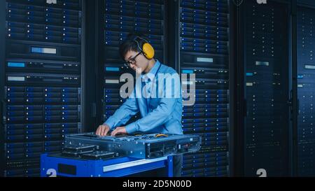 In the Modern Data Center: IT Technician Wearing Protective Headphones Working with Server Racks, on a Pushcart Installing New Hardware. Engineer Stock Photo