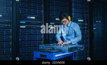 In the Modern Data Center: IT Technician Wearing Protective Headphones Working with Server Racks, on a Pushcart Installing New Hardware. Engineer Stock Photo