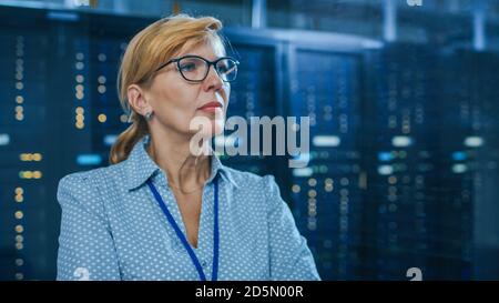 Portrait of a Beautiful Senior Female IT Technician Standing In Modern Data Center. In the Background Working Server Racks with Blinking LED Lights. Stock Photo