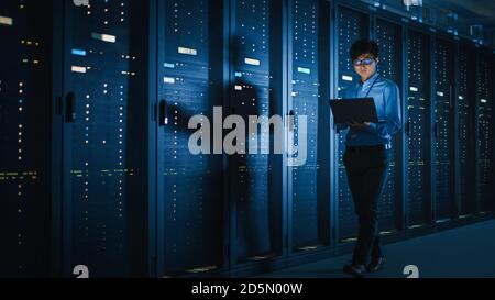 In Dark Data Center: Male IT Specialist Walks along the Row of Operational Server Racks, Uses Laptop for Maintenance. Concept for Cloud Computing Stock Photo