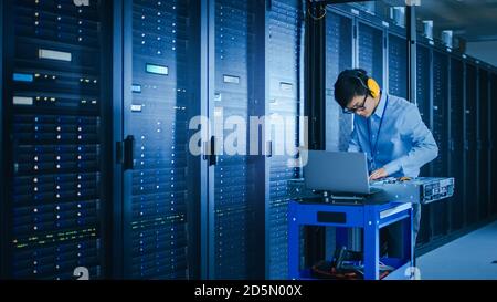 In the Modern Data Center: IT Technician Wearing Protective Headphones Working with Server Racks, on a Pushcart Installing New Hardware. Engineer Stock Photo