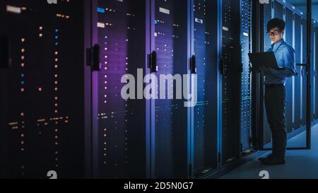 In Dark Data Center: Male IT Specialist Stands Beside the Row of Operational Server Racks, Uses Laptop for Maintenance. Concept for Cloud Computing Stock Photo