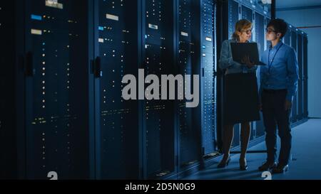 In the Modern Data Center: Engineer and IT Specialist Work with Server Racks, Talking, Doing System Maintenance Check with Laptop Computer. Concept of Stock Photo