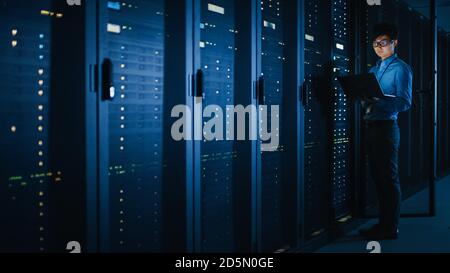 In Dark Data Center: Male IT Specialist Stands Beside the Row of Operational Server Racks, Uses Laptop for Maintenance. Concept for Cloud Computing Stock Photo