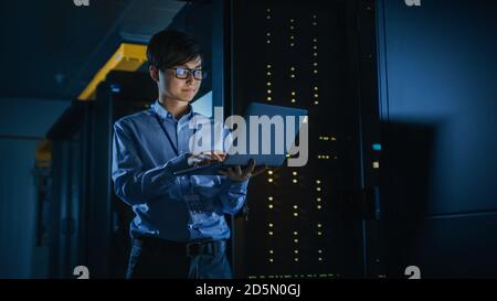 In Dark Data Center: Male IT Specialist Stands Beside the Row of Operational Server Racks, Uses Laptop for Maintenance. Concept for Cloud Computing Stock Photo