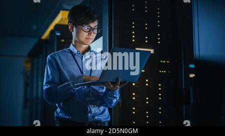 In Dark Data Center: Male IT Specialist Stands Beside the Row of Operational Server Racks, Uses Laptop for Maintenance. Concept for Cloud Computing Stock Photo