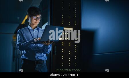 In Dark Data Center: Male IT Specialist Stands Beside the Row of Operational Server Racks, Uses Laptop for Maintenance. Concept for Cloud Computing Stock Photo