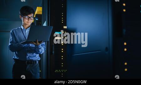 In Dark Data Center: Male IT Specialist Stands Beside the Row of Operational Server Racks, Uses Laptop for Maintenance. Concept for Cloud Computing Stock Photo