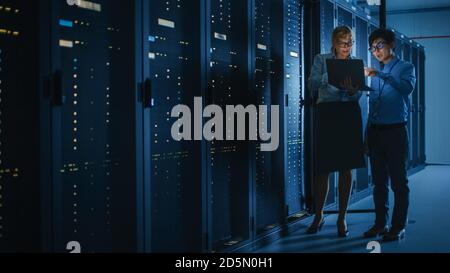 In the Modern Data Center: Engineer and IT Specialist Work with Server Racks, Talking, Doing System Maintenance Check with Laptop Computer. Concept of Stock Photo