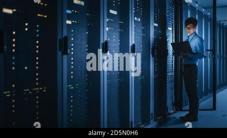 In Dark Data Center: Male IT Specialist Stands Beside the Row of Operational Server Racks, Uses Laptop for Maintenance. Concept for Cloud Computing Stock Photo