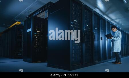 In Data Center: Male IT Specialist Wearing White Coat Stands Beside Server Rack, Uses Laptop Computer to Run Maintenance Diagnostics Tools, Controls Stock Photo