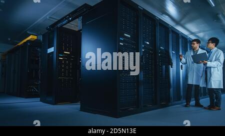 In the Data Center: Two IT Specialists Wearing White Lab Coats, Have Discussion about Working of Server Racks. Optimizing Work of Database. Cloud Stock Photo