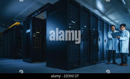 In the Data Center: Two IT Specialists Wearing White Lab Coats, Have Discussion about Working of Server Racks. Optimizing Work of Database. Cloud Stock Photo