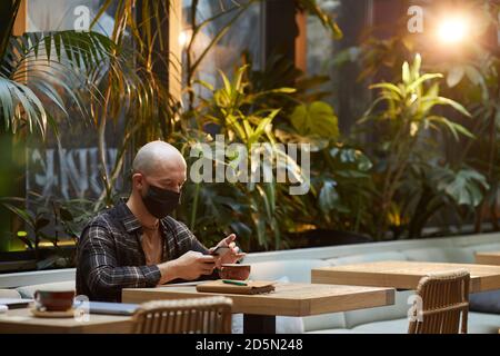 Young man in protective mask using mobile phone while sitting at the table in cafe Stock Photo