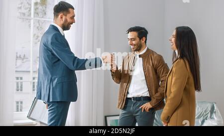 Professional Real Estate Agent Shows Bright New Apartment to a Young Couple. Successful Young Couple Becoming Homeowners are Getting Keys From New Stock Photo