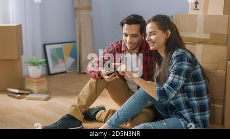 Happy Young Couple Sitting on the Floor of the Newly Rented / Purchased Apartment Use Smartphone. Unpacked Cardboard Boxes and Covered Furniture in Stock Photo