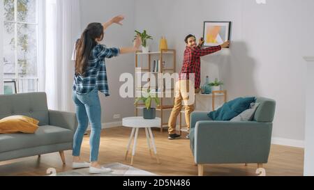 Happy Couple Hanging Picture on the Wall, Boyfriend Moves It, Girlfriend Tells Him when the Frame is Hanging Straight. Funny Moment in Young Couple's Stock Photo