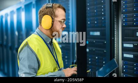 Bearded IT Specialist in Glasses and Headphones, wearing High Visibility Vest is Working on Laptop in Data Center Next to Server Racks. Running Stock Photo