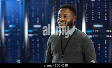 Black Data Center IT Technician Standing in the Middle of a Server Rack Corridor. He is Smiling. Stock Photo