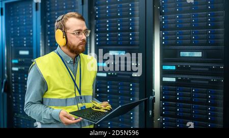 Bearded IT Specialist in Glasses and Headphones, wearing High Visibility Vest is Working on Laptop in Data Center Next to Server Racks. Running Stock Photo