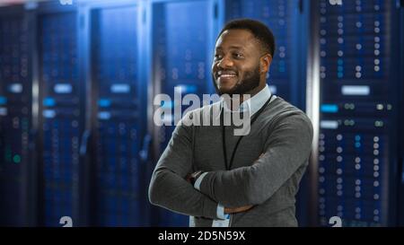 Black Data Center IT Technician Standing in the Middle of a Server Rack Corridor. He is Standing with Crossed Arms and is Smiling. Stock Photo