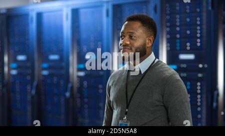 Black Data Center IT Technician Standing in the Middle of a Server Rack Corridor. He is Smiling. Stock Photo
