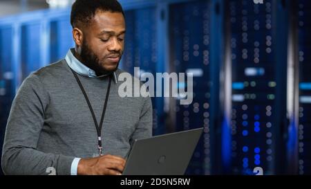 Black Data Center IT Technician Standing in Server Rack Corridor with a Laptop Computer. He is Visually Inspecting Working Server Cabinets. Stock Photo